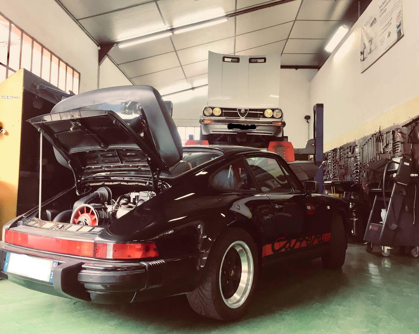 Dans l'atelier : une Porsche 911 noire capot ouvert, une Alfa Romeo sur le pont, établi et outillage au mur