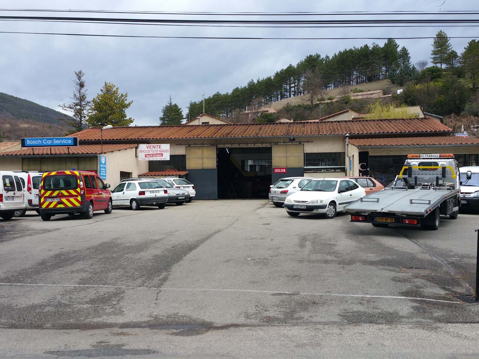 Façade du Garage des Trois Becs à Saillans : atelier au toit de tuiles, voitures sur le parking, collines de pins derrière
